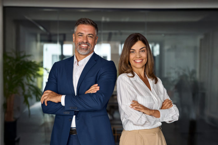 Vertical portrait of smiling mature Latin hispanic business man and European business woman standing arms crossed in office. Diverse colleagues, group team of confident professional business people.